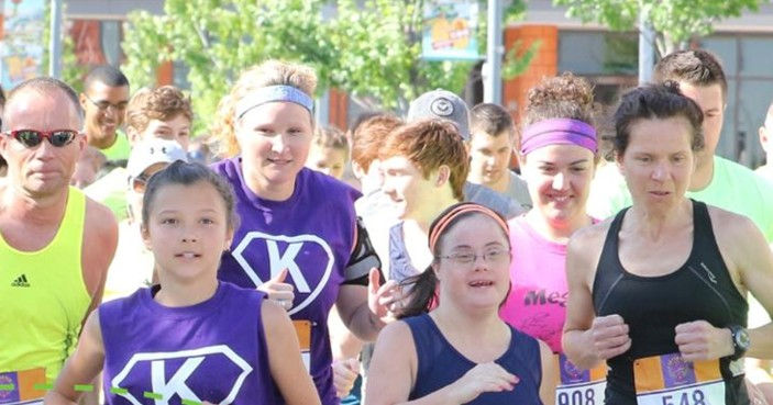 Group of runners race outdoors in bright sun, including women in purple K shirts and bibs, with focused expressions.