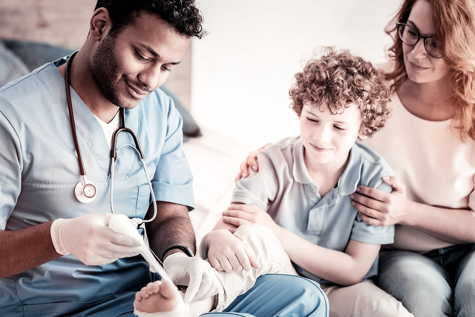 A nurse in blue scrubs gently bandages a boy's foot while a woman, possibly his mother, comforts him. The setting is calm and supportive.