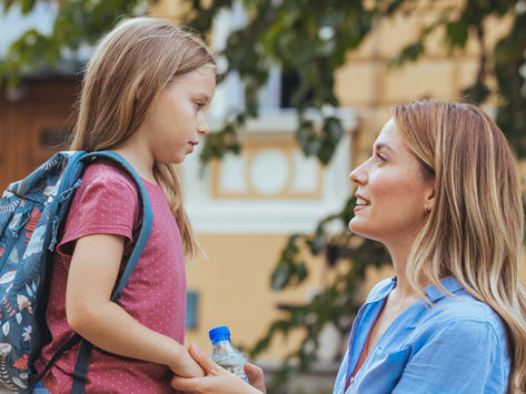 Mother and young daughter hold hands outdoors, smiling and talking; girl wears a backpack and mom holds a water bottle.