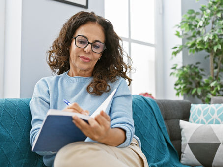 Woman in blue sweater writing in a notebook on a sofa. Background features green plant and cushions with geometric patterns, soft light.