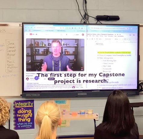 A classroom with students facing a large screen showing a man speaking. A boy points at the screen. Text on screen: "The first step for my Capstone project is research." Whiteboard background with notes. Navy and yellow colors dominate. Mood is focused.