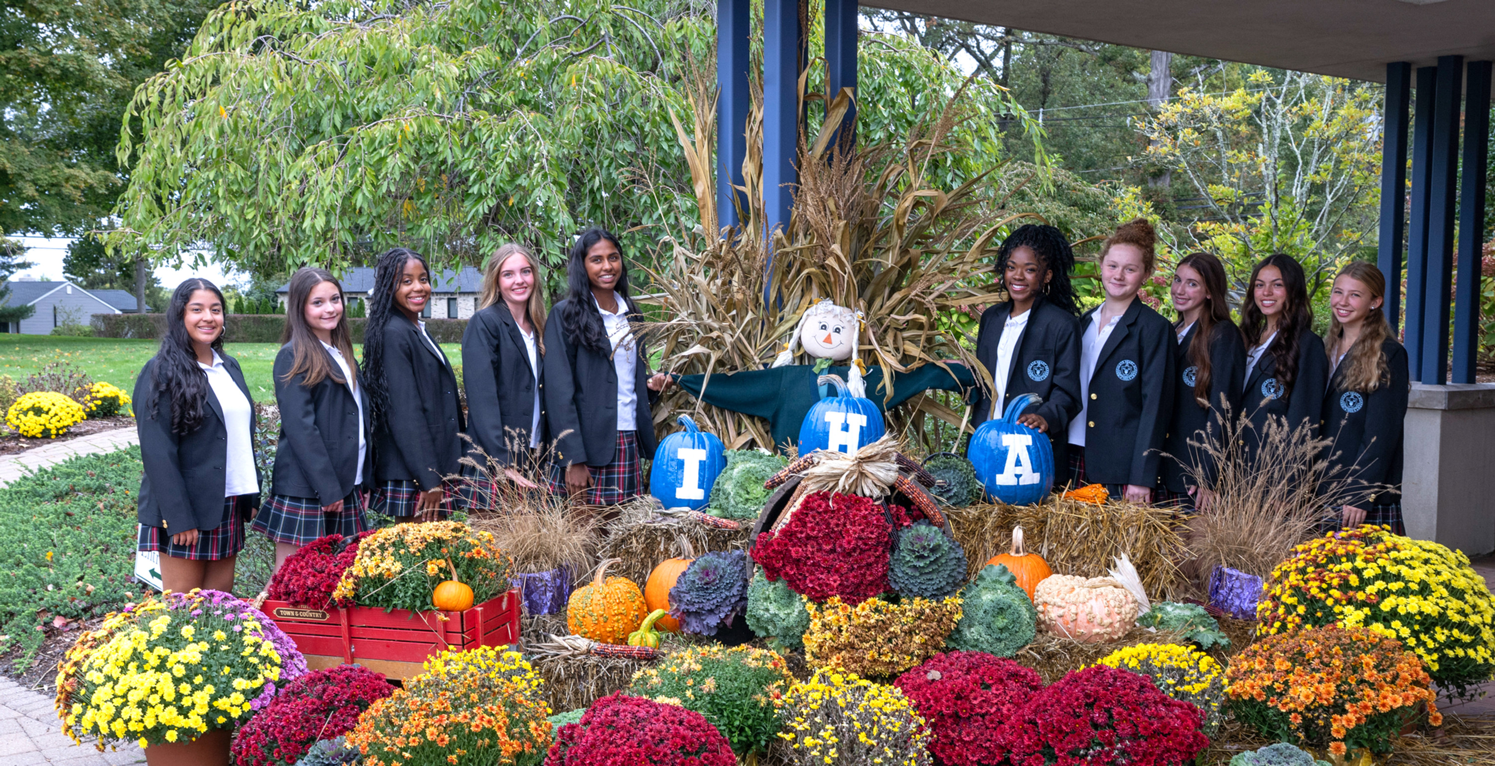 Smiling schoolgirls in blazers pose beside a fall display of flowers, pumpkins and hay, with blue pumpkins spelling T H A.