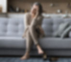 Woman sitting on grey sofa, looking thoughtful with head in hand. She is barefoot, shoes on floor, kitchen in background. Neutral tones.
