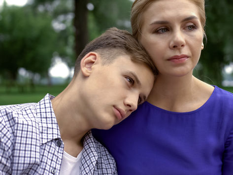 Woman in purple shirt hugs a boy in a checkered shirt as he rests his head on her shoulder in a green park.