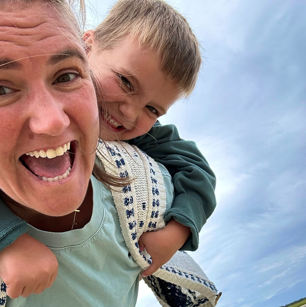 Woman carrying a smiling child on her back. Both are laughing. The child is wearing a green shirt. Blue sky in the background.