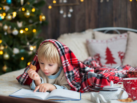 Child under a red plaid blanket reads a book, holding a cookie. Background features a festive Christmas tree with warm lights. Cozy mood.