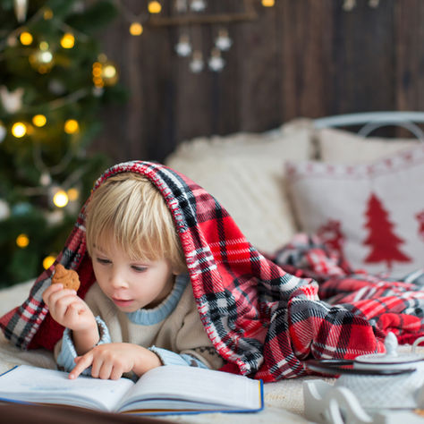 Child under a red plaid blanket reads a book, holding a cookie. Background features a festive Christmas tree with warm lights. Cozy mood.