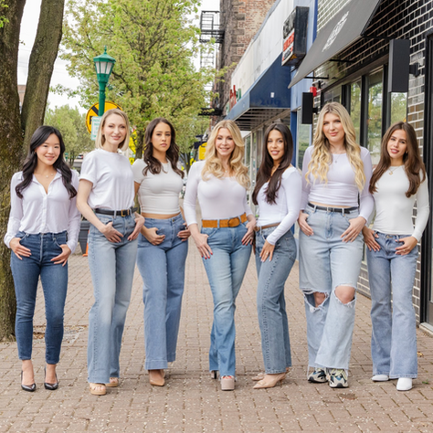 Seven women in white tops and jeans stand on a sidewalk, smiling. Trees and shops line the background, creating a casual, cheerful mood.