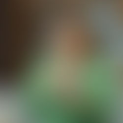 Woman in a green shirt looking concerned while reading her phone, sitting on a gray sofa with a wooden shelf background.