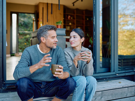 Couple sits on wooden porch steps, chatting and holding mugs outside a modern home, with warm autumn light.