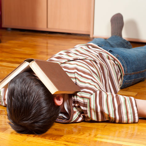 Child lying face down on wooden floor with book on head, wearing striped shirt and jeans, in a room with cabinets, looking exhausted. Bergen County Moms