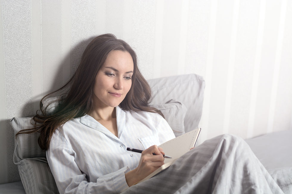 Woman in pajamas writes in a notebook while sitting on a bed. Soft lighting, white and gray striped background, calm expression.