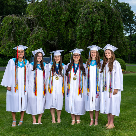 Six smiling female graduates in white caps and gowns stand on a green lawn with leafy trees behind them.