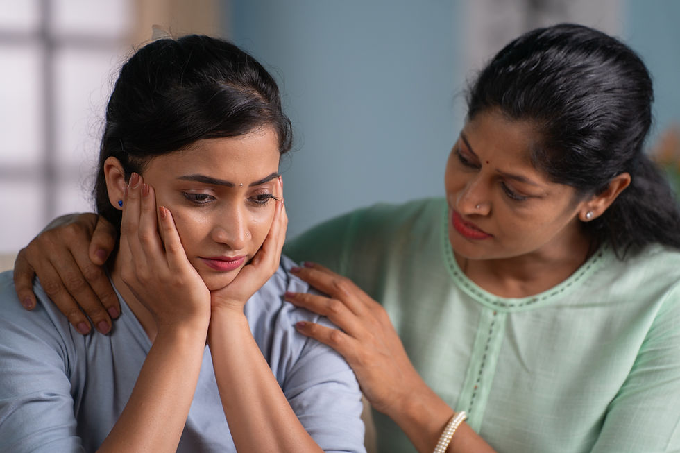Two women indoors, one comforts a sad younger woman holding her face, with a reassuring hand on her shoulder.