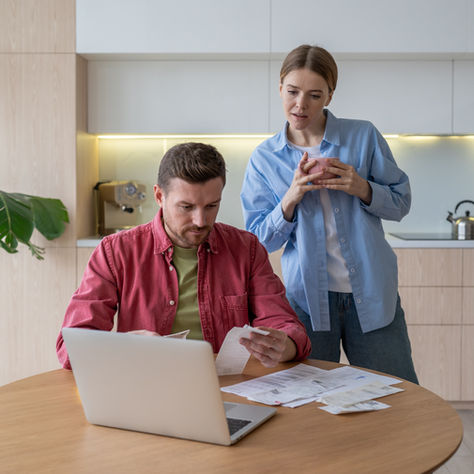 Man and woman review bills at a laptop in a bright modern kitchen, looking focused and concerned, with a plant and kettle in back
