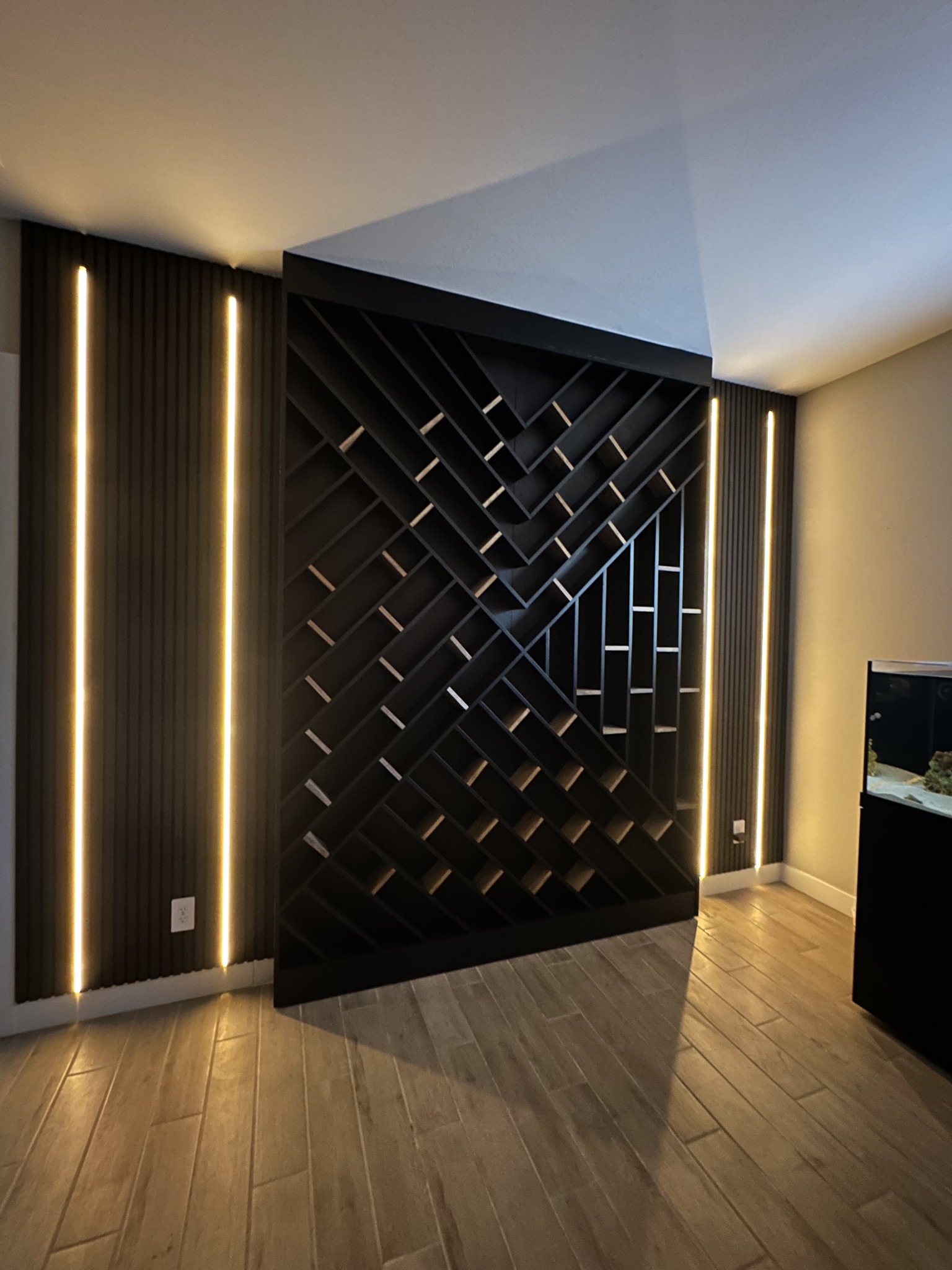 Black acoustic slat wood wall panels framing a geometric room divider with built-in shelving and warm vertical lights