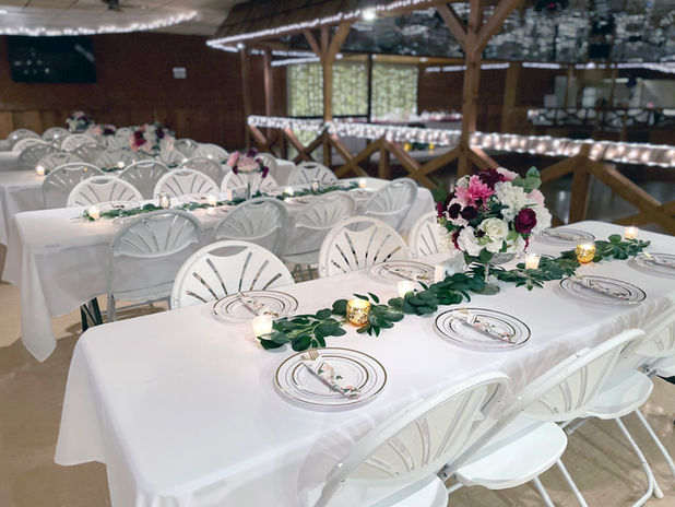 Several banquet tables covered in white linens with greenery, candles, and a floral mercury bowl centerpiece are surrounded by white plastic chairs. There is a sunken wooden dance floor with mirrored ceiling and a lighted organza backdrop in the background.