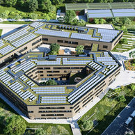 Luftaufnahme eines großen, modernen Schulgebäudes mit begrüntem Dach und Sonnenkollektoren, umgeben von üppigem Grün, Straßen mit Autos und einer ruhigen Landschaft mit Bäumen und Rasenflächen.