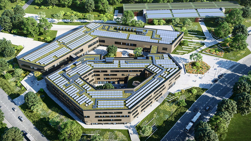 Luftaufnahme eines großen, modernen Schulgebäudes mit begrüntem Dach und Sonnenkollektoren, umgeben von üppigem Grün, Straßen mit Autos und einer ruhigen Landschaft mit Bäumen und Rasenflächen.