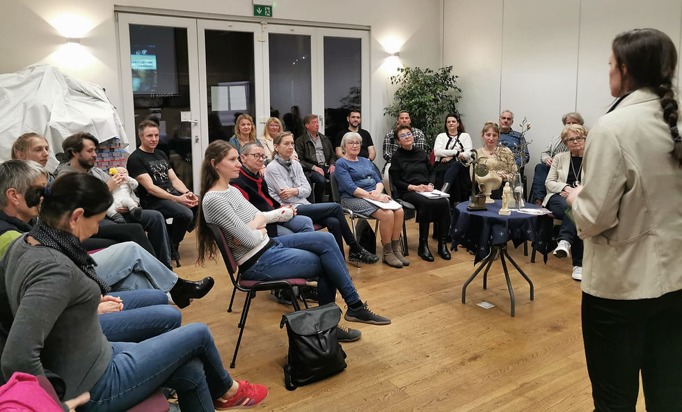 Audience listens to a woman speaking in a bright room, with small sculptures on a table and an exit sign above the doors.