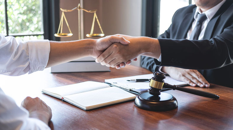 Two people in suits shake hands over a table with a gavel and open notebook. Scales of justice in the background. Professional setting.