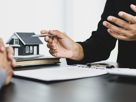 Hands discussing real estate over a contract with a miniature house model on a table. Formal setting, neutral tones, focused negotiation.