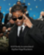 A$AP Rocky in black suit and sunglasses poses on a floral-themed red carpet. Background shows people and cameras. Confident expression.