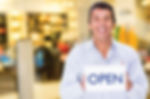 A smiling business owner holding an open sign in front of a store.