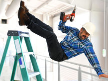 Worker in a blue plaid shirt falls off a green ladder in a bright warehouse, holding a drill. He wears a hard hat, showing a surprised expression.