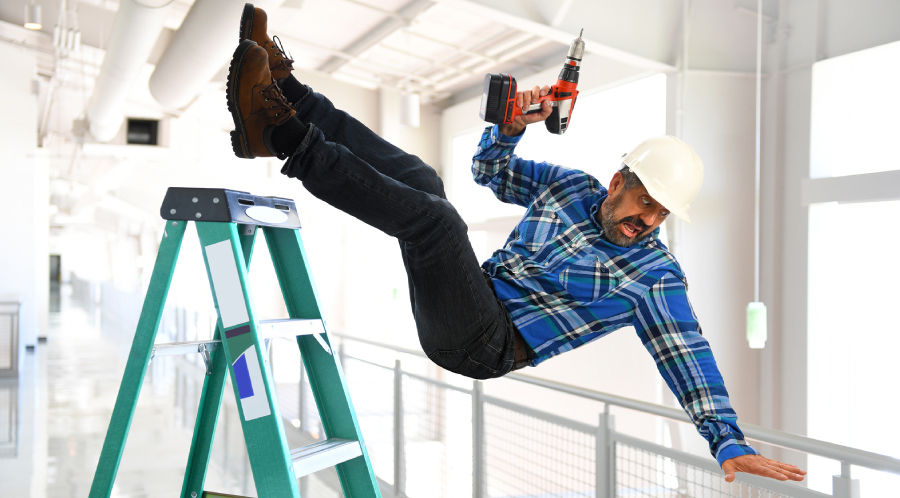 Worker in a blue plaid shirt falls off a green ladder in a bright warehouse, holding a drill. He wears a hard hat, showing a surprised expression.