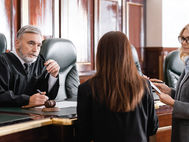 A judge in a courtroom listens to a lawyer presenting documents. Gavel on desk. The setting is formal with dark wood paneling.