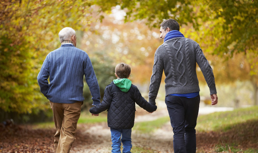 Three people, a child between two adults, walk hand-in-hand on a leafy autumn path. They wear sweaters; trees display fall colors.