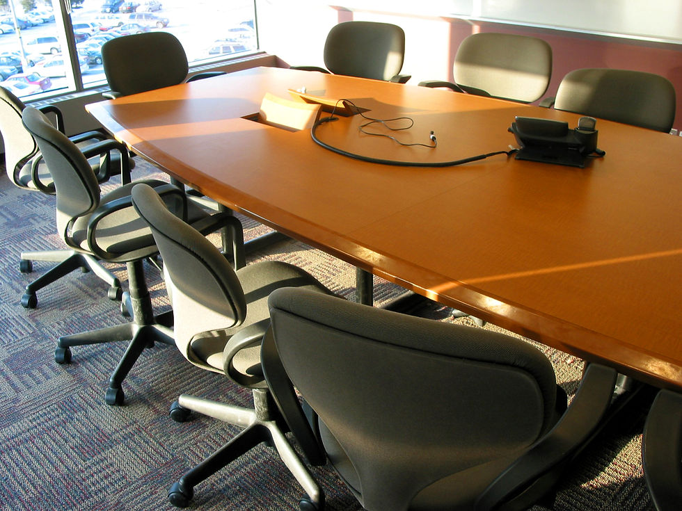 Conference room with a wooden table, gray chairs, phone, and cables. Sunlight streams through large windows, adding warmth to the setting.