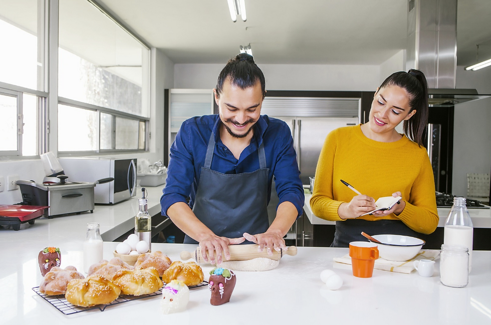 Two people in a bright kitchen; a man in a dark apron rolls dough, smiling, while a woman in a yellow sweater takes notes. Baked bread nearby.
