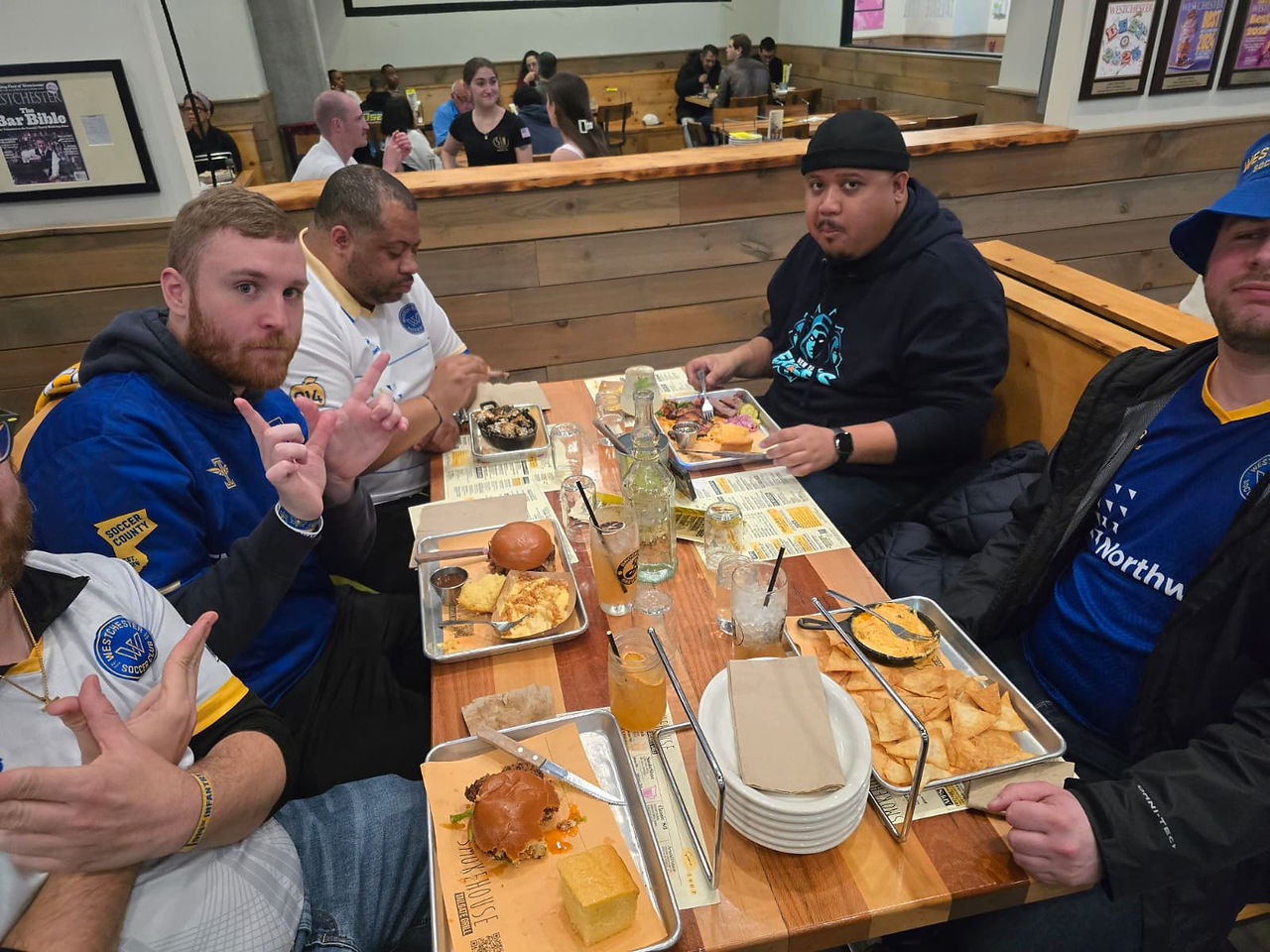 A group of Westchester SC supporters wearing blue and white club gear gather around a wooden table at Smokehouse Tailgate Grill in New Rochelle after the match. Plates of BBQ, burgers, chips, and drinks cover the table as fans pose for the camera inside the busy restaurant.