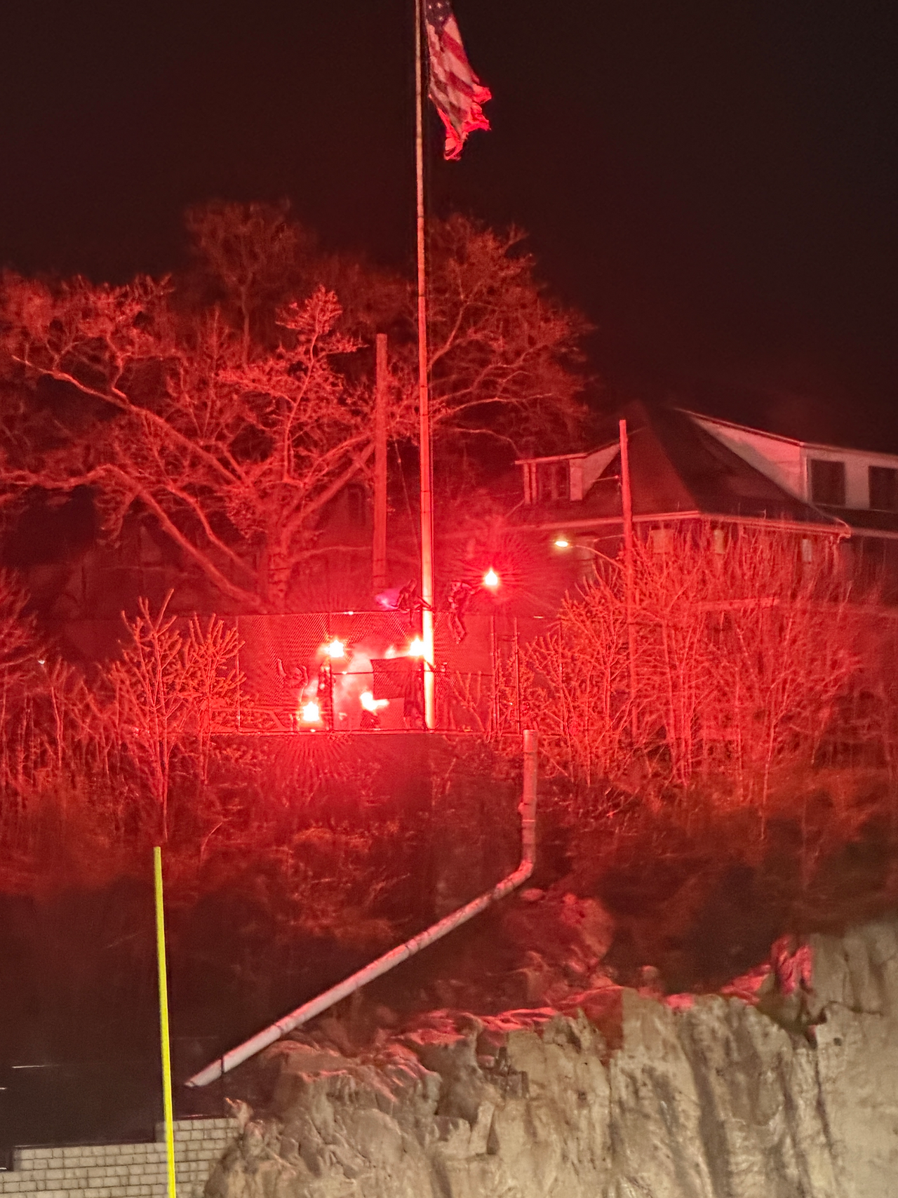 Bright red flares burn along a fence line near a flagpole and nearby houses, casting a red glow over trees and the surrounding area at night.