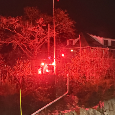 Bright red flares burn along a fence line near a flagpole and nearby houses, casting a red glow over trees and the surrounding area at night.