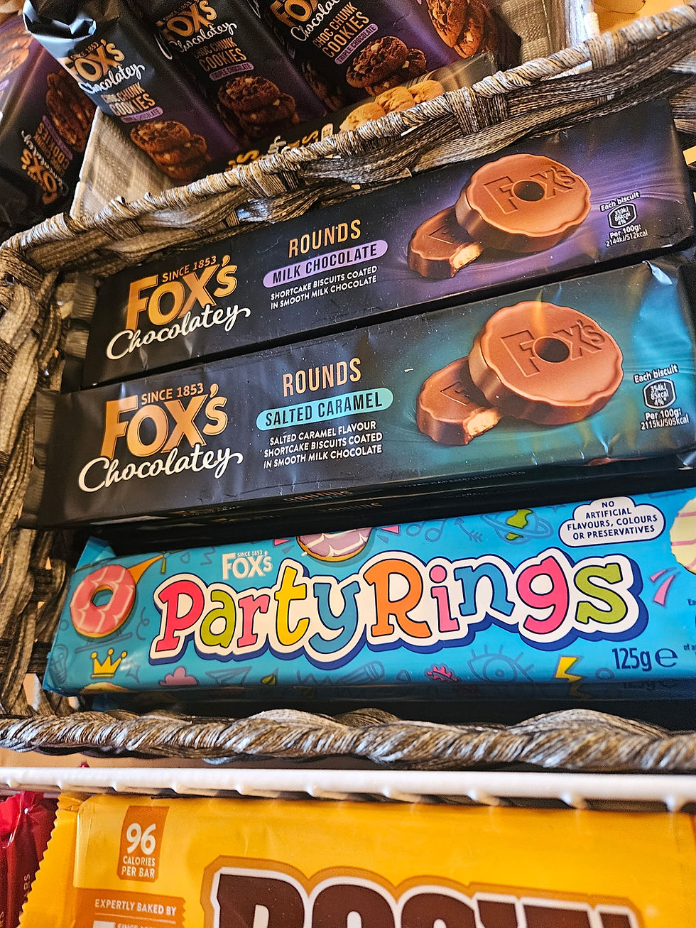 A basket display of British snacks featuring Fox’s Chocolatey biscuit rounds in milk chocolate and salted caramel varieties, along with colorful Party Rings, stacked neatly in a shop setting.