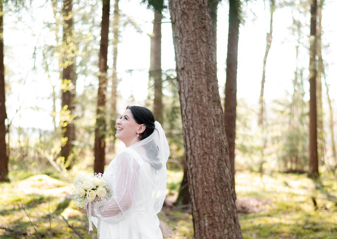 Braut im Wald, lächelnd mit Brautstrauß, HOCHZEITSFOTOGRAFIE, von Bäumen umgeben.