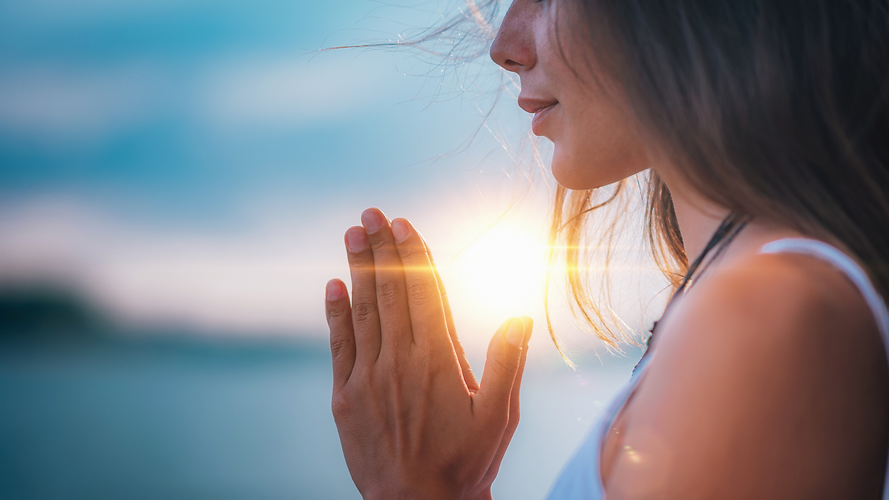 Woman with her hands put together in a praying form, with the sun shining from behind her hands,