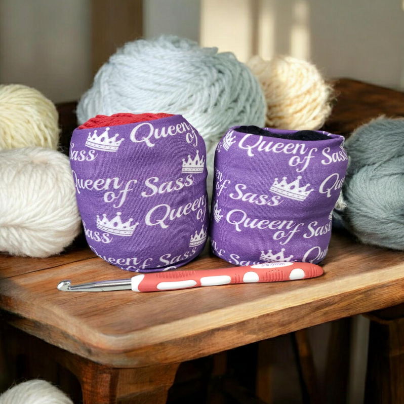 yarn balls, crochet hook on wooden table with yarn cozies that say queen of sass