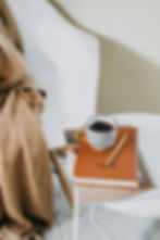 Cozy scene with a striped mug of coffee on a table, atop a brown journal and magazines. A beige blanket drapes over a white chair. Relaxing mood.