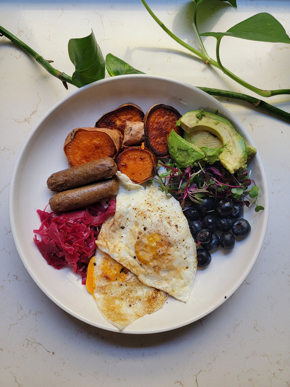 Healthy balanced breakfast bowl with eggs, sweet potatoes, chicken sausage, avocado, blueberries, microgreens, and fermented red cabbage arranged on a white plate.