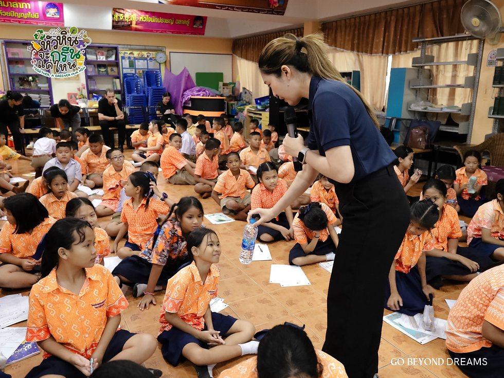 ครูถือไมโครโฟนกำลังมอบขวดน้ำให้กับนักเรียนที่นั่งอยู่ภายในห้องเรียน โดยนักเรียนสวมชุดสีส้ม