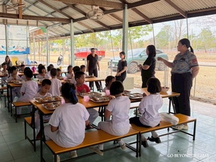 นักเรียนนั่งรับประทานอาหารกลางวันที่โต๊ะในโรงอาหาร โดยมีคุณครูยืนอยู่ใกล้ๆ