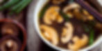 Bowl of bone broth mushroom soup with green onions, chopsticks on top, wooden table background. Shiitake mushrooms and scallions on the side.