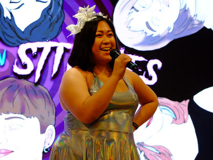 A comedian stands on stage holding a microphone and smiling mid-performance. She wears a metallic silver dress, sneakers, and a decorative headpiece. Behind her, a large screen displays stylized illustrated portraits and the words “Bitches in Stitches,” the name of the feminist comedy collective.