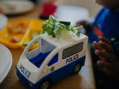 A child’s dark-skinned hand reaches toward a blue-and-white toy police van on a wooden table, with bits of food and a child’s plate visible in the background.