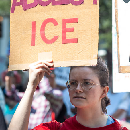 A woman wearing glasses and a red shirt holds a cardboard protest sign reading “Abolish ICE” above their head during a demonstration. Other protesters and signs are visible blurred in the background.