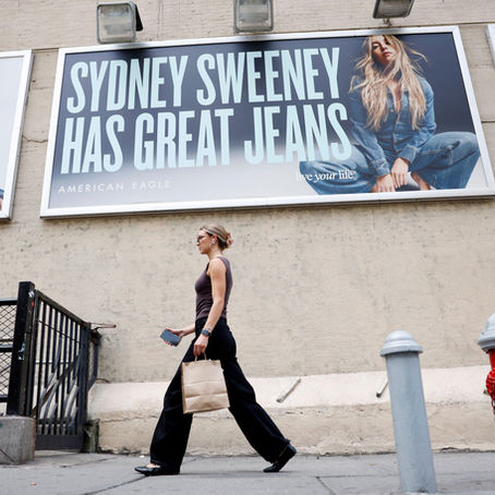 A person walks past large outdoor billboards for American Eagle featuring model Sydney Sweeney in denim outfits. The main sign reads “SYDNEY SWEENEY HAS GREAT JEANS.” The person in the foreground is wearing sunglasses, black pants, a dark tank top, and carrying a shopping bag and phone while passing a red fire hydrant on the sidewalk.